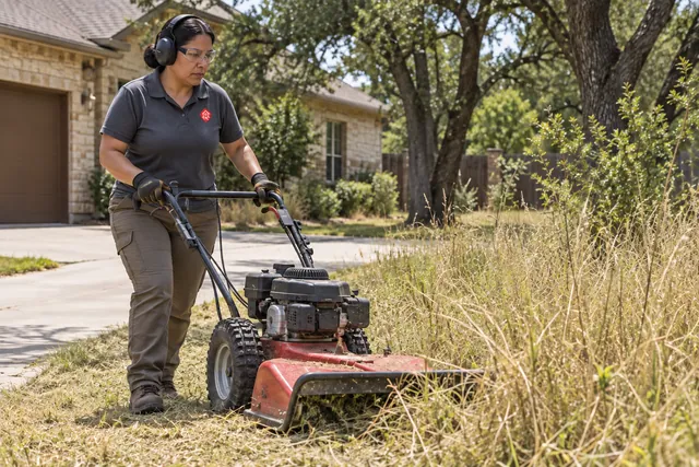 Worker clearing tall grass beside a driveway with a wheeled brush mower